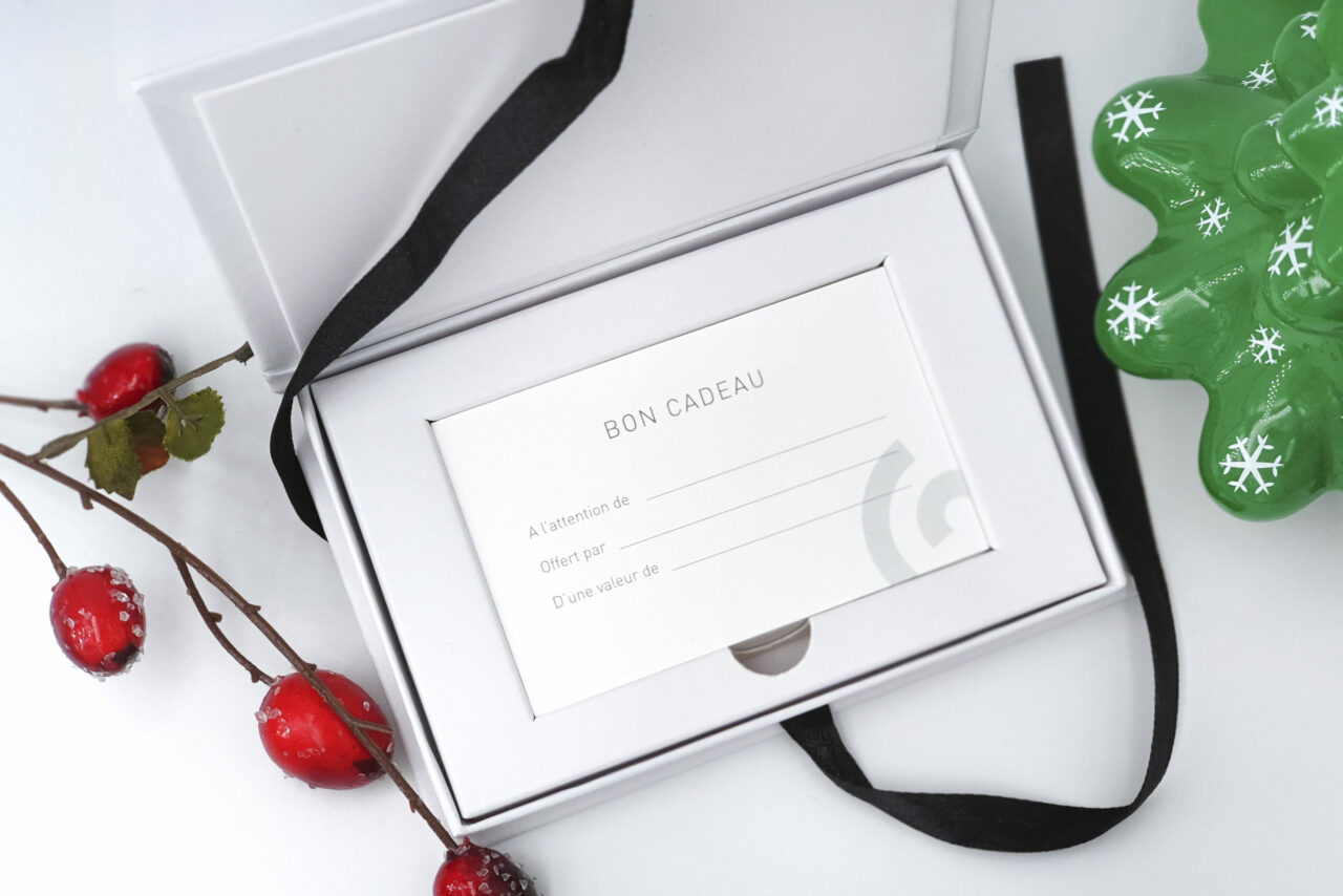 An open gift box containing a blank gift card labeled Bon Cadeau in French. The box includes black ribbon details and is placed next to decorative red berries and a green fir-shaped ornament with snowflake designs.