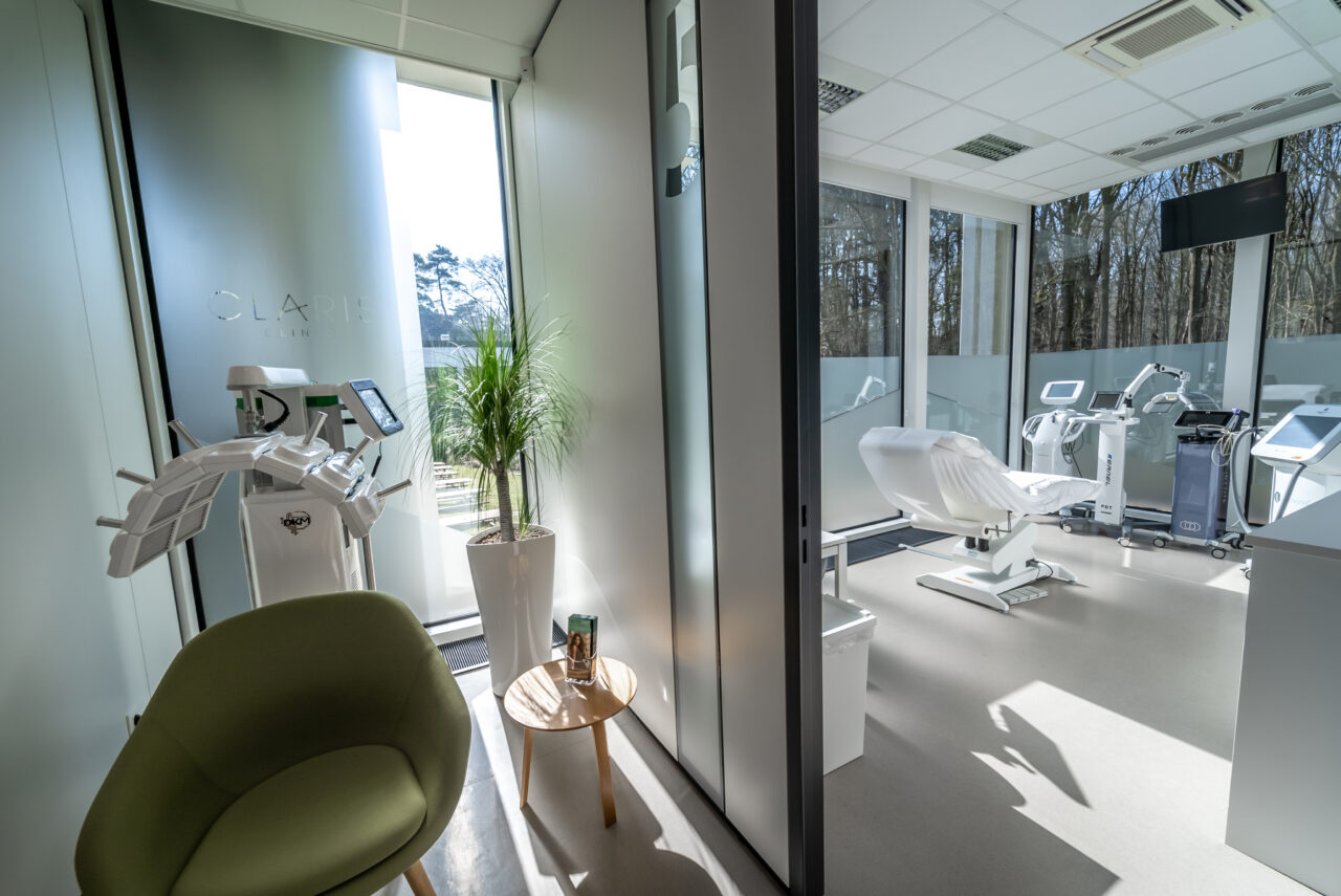 Cabinet dentaire moderne avec une chaise verte et une petite table ronde au premier plan. Une plante en pot trône près des parois vitrées. Du matériel dentaire et une chaise sont visibles dans la salle de soins lumineuse en arrière-plan. De grandes baies vitrées offrent une vue sur la forêt.