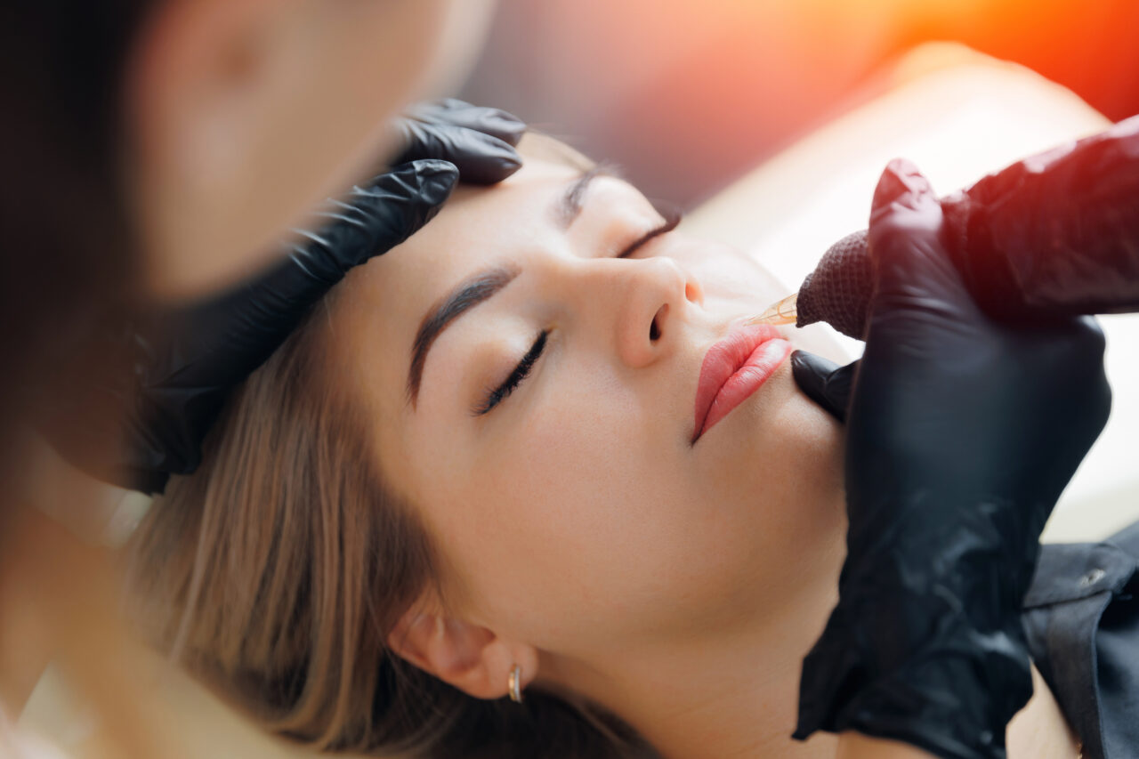 Une personne portant des gants noirs réalise un tatouage cosmétique ou un maquillage permanent sur les lèvres d'une autre personne, allongée, les yeux fermés. La scène est très éclairée, avec une douce lueur orange en arrière-plan.