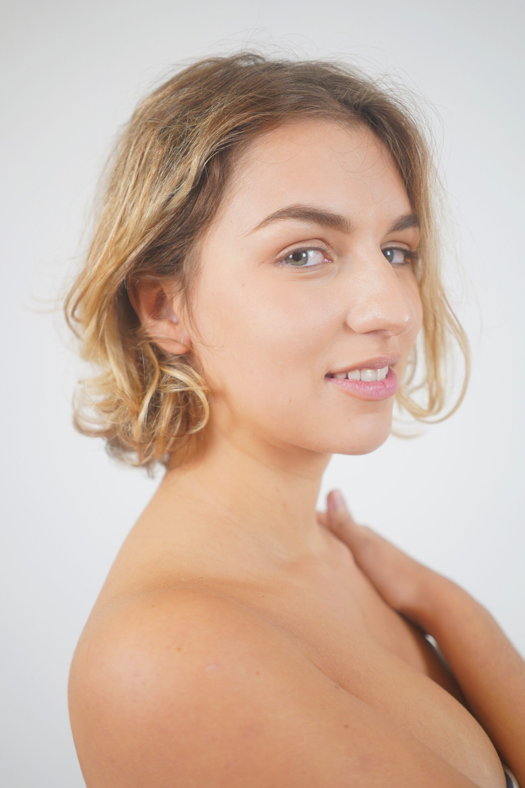 Une personne aux cheveux blonds courts et ondulés regarde vers l'objectif avec un léger sourire. Elle a la peau claire et se tient sur un fond blanc uni.