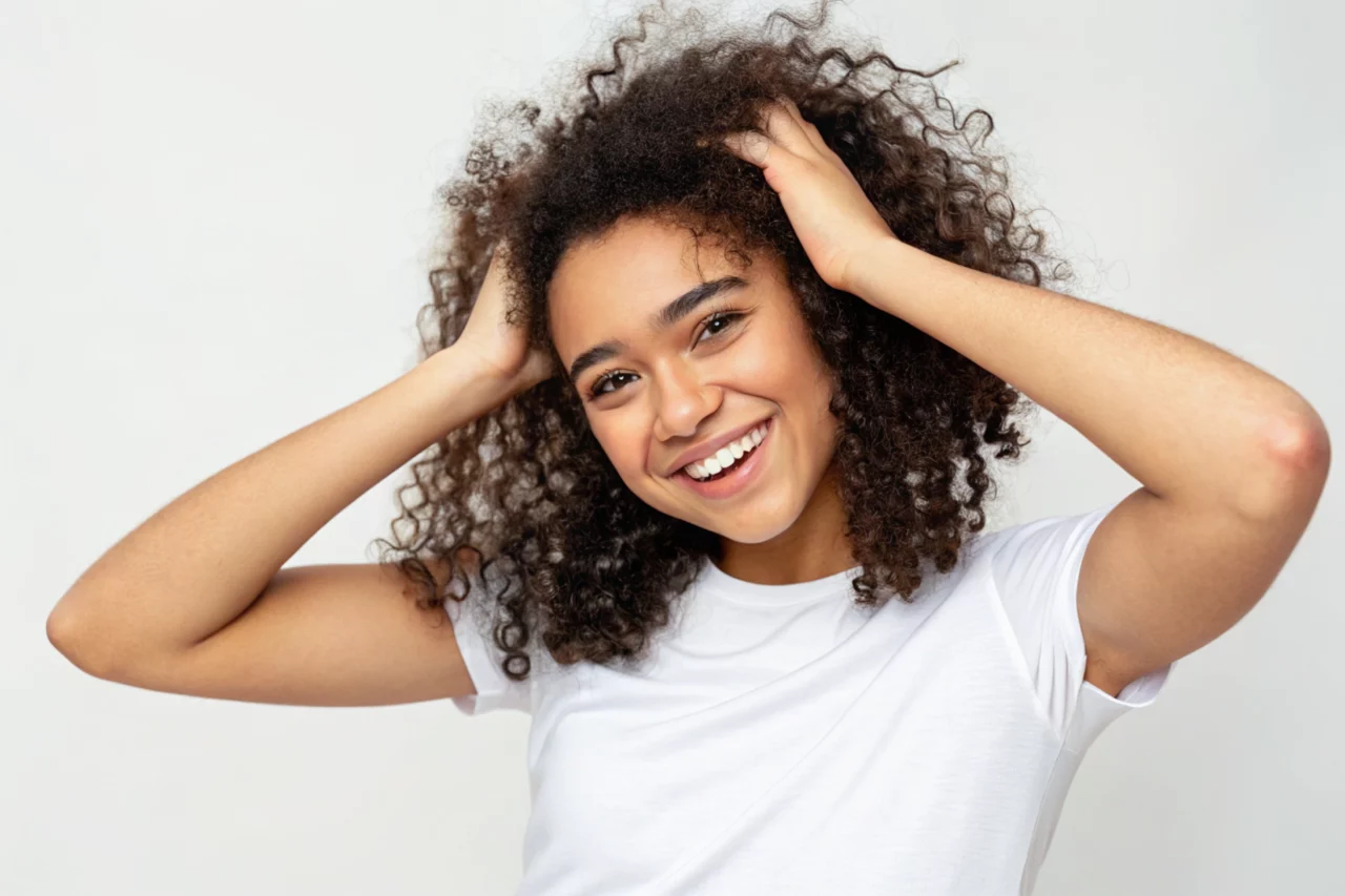 Une personne aux cheveux bouclés et portant un t-shirt blanc sourit tout en portant ses mains à sa tête sur un fond uni.
