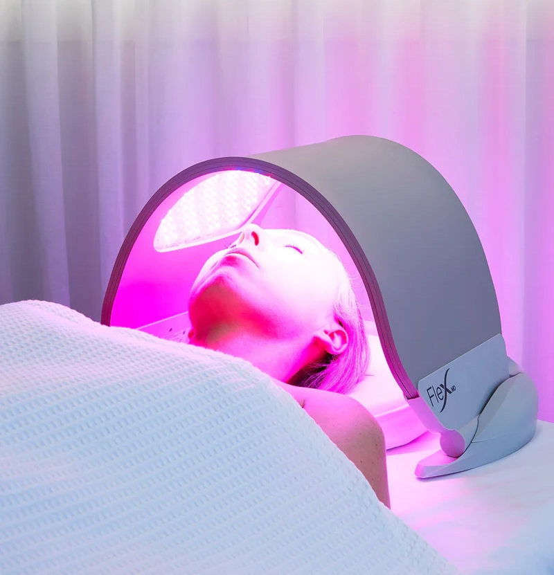 Une personne est allongée sous un appareil incurvé émettant une lumière rose, recevant peut-être une luminothérapie LED. Elle est recouverte d'une couverture blanche et entourée de rideaux blancs.