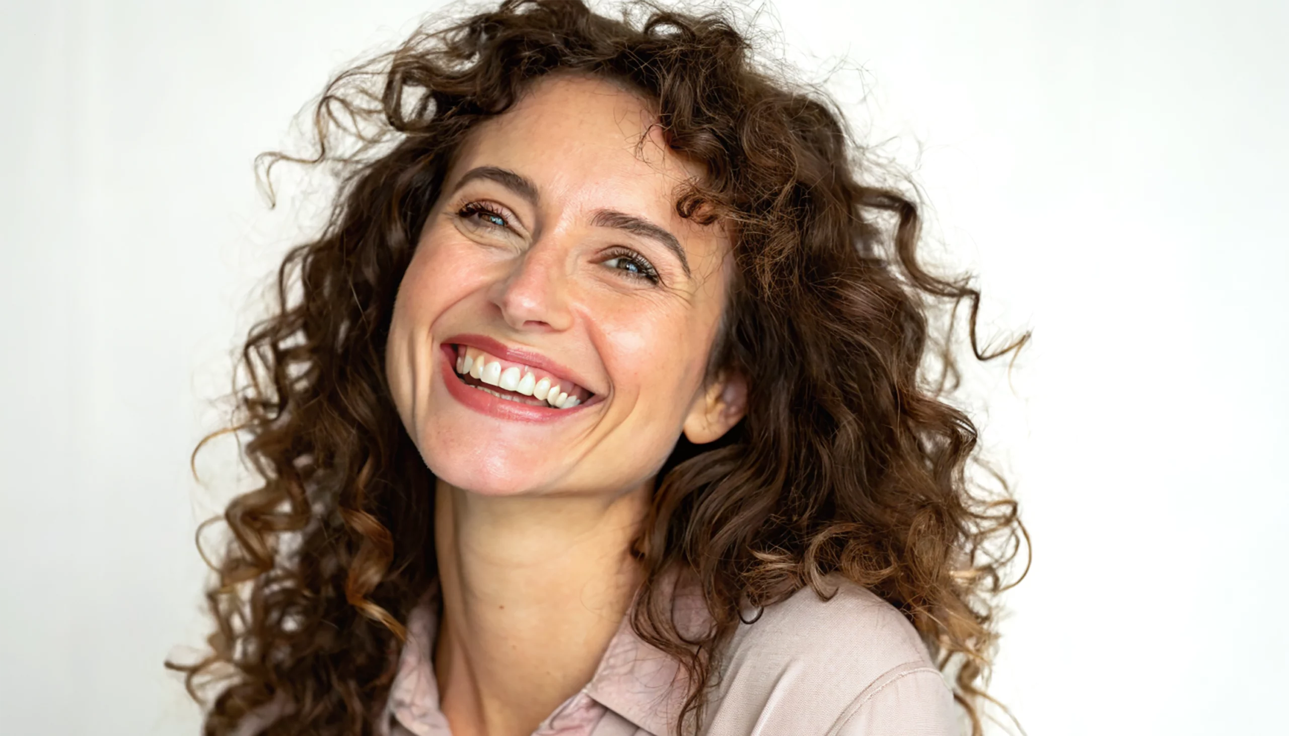 Une personne aux cheveux bouclés arbore un large sourire, le regard légèrement levé. Elle porte une chemise claire et se détache sur un fond blanc uni.