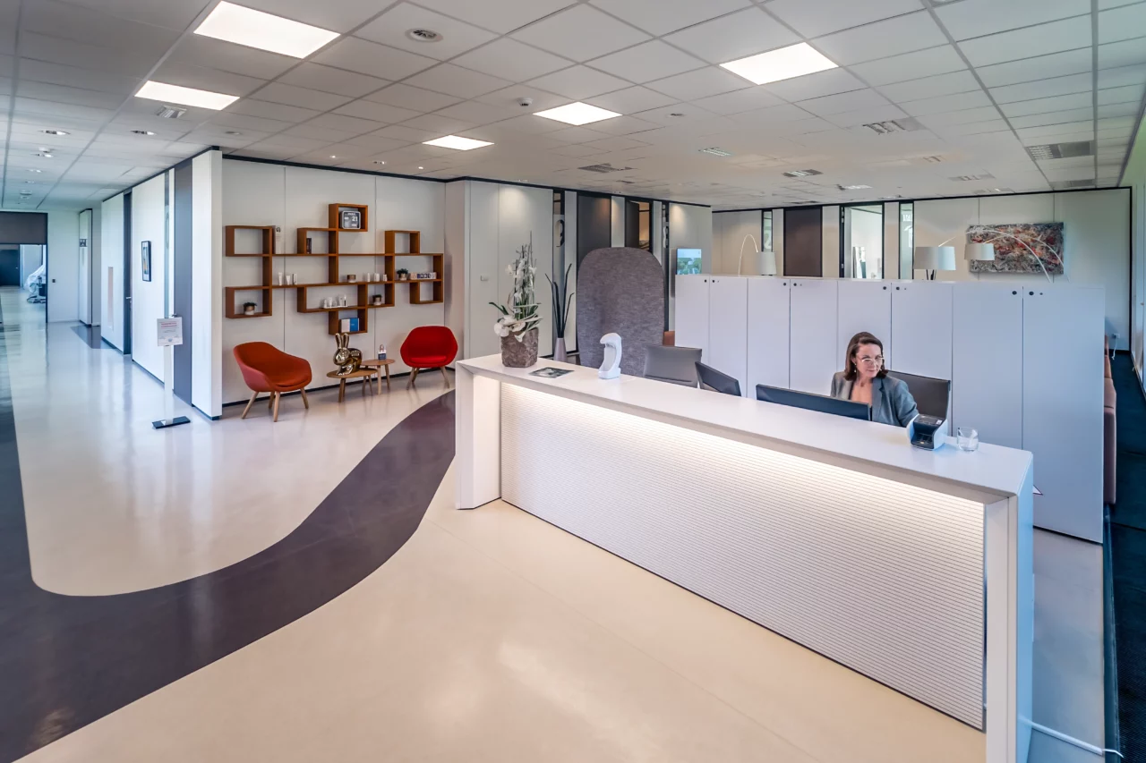 Un espace de réception moderne avec un bureau blanc et une femme au téléphone au comptoir. Derrière elle, des placards et un coin salon ouvert avec deux chaises et une petite table. La pièce est ornée de plafonniers et d'un sol au motif géométrique.