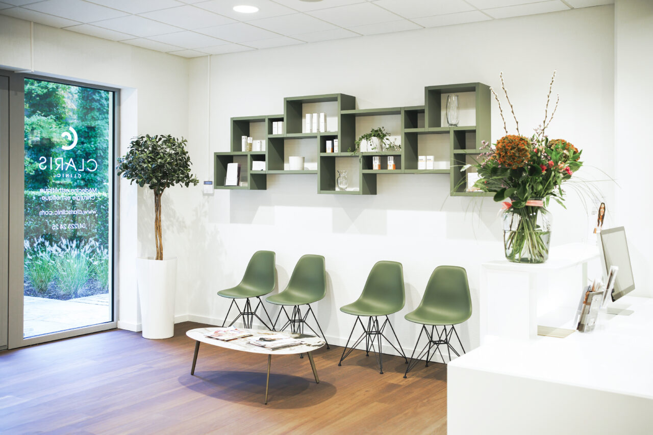 Une salle d'attente moderne avec quatre chaises vertes et une table basse en bois sur laquelle sont posés des magazines. Un mur est orné d'étagères géométriques ornées d'objets et de produits décoratifs. Une grande plante en pot est placée près de la porte, marquée « Claris ». Un vase de fleurs est posé sur le bureau.