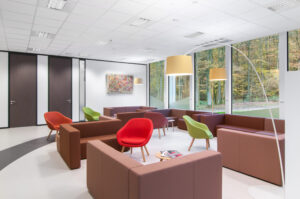 Une salle d'attente moderne avec des canapés marron, des chaises rouges et vertes, deux grands lampadaires et un tableau abstrait coloré au mur. De grandes fenêtres offrent une vue sur la forêt. Quelques magazines sont posés sur l'une des tables.