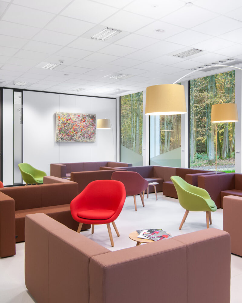 Une salle d'attente moderne avec des canapés marron, des chaises rouges et vertes, deux grands lampadaires et un tableau abstrait coloré au mur. De grandes fenêtres offrent une vue sur la forêt. Quelques magazines sont posés sur l'une des tables.