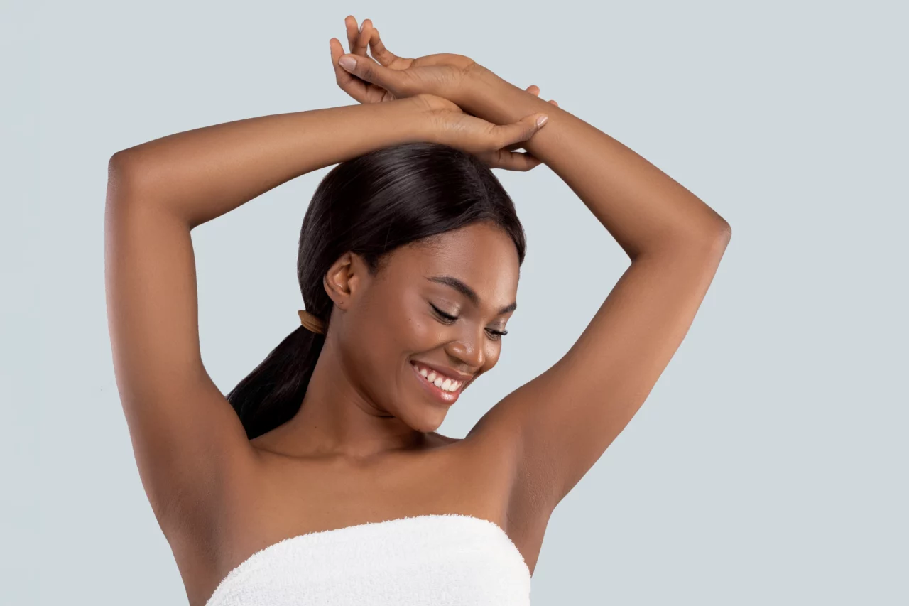 Une femme aux cheveux longs attachés en queue de cheval sourit, les yeux fermés. Elle a les bras levés et croisés au-dessus de la tête. Elle porte une serviette blanche et se tient debout sur un fond gris clair uni.