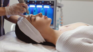 Une personne allongée sur un lit de traitement reçoit d'un praticien ganté un soin du visage à l'aide d'un appareil manuel. La personne porte un bandeau et un peignoir blancs, et le cadre semble être celui d'une clinique médicale ou d'une station thermale.