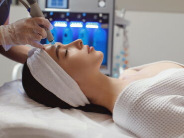 Une personne allongée sur un lit de traitement reçoit d'un praticien ganté un soin du visage à l'aide d'un appareil manuel. La personne porte un bandeau et un peignoir blancs, et le cadre semble être celui d'une clinique médicale ou d'une station thermale.