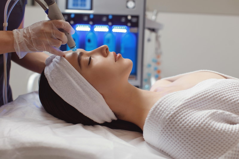 Une personne allongée sur un lit de traitement reçoit d'un praticien ganté un soin du visage à l'aide d'un appareil manuel. La personne porte un bandeau et un peignoir blancs, et le cadre semble être celui d'une clinique médicale ou d'une station thermale.