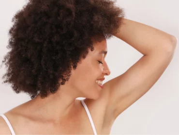 Une personne aux cheveux bouclés sourit, les yeux fermés, en levant un bras et en penchant la tête en arrière. Elle porte un débardeur blanc et se tient debout sur un fond clair uni.