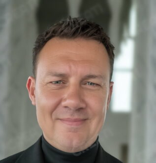 Un homme aux cheveux bruns courts, aux yeux clairs et au léger sourire porte un col roulé noir. L'arrière-plan est flou et laisse apparaître de grandes fenêtres et des détails architecturaux blancs.