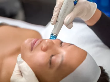 A person lies on a treatment bed with eyes closed while a gloved professional uses a HydraFacial device on their cheek. The person wears a white headband and appears relaxed during the facial procedure.