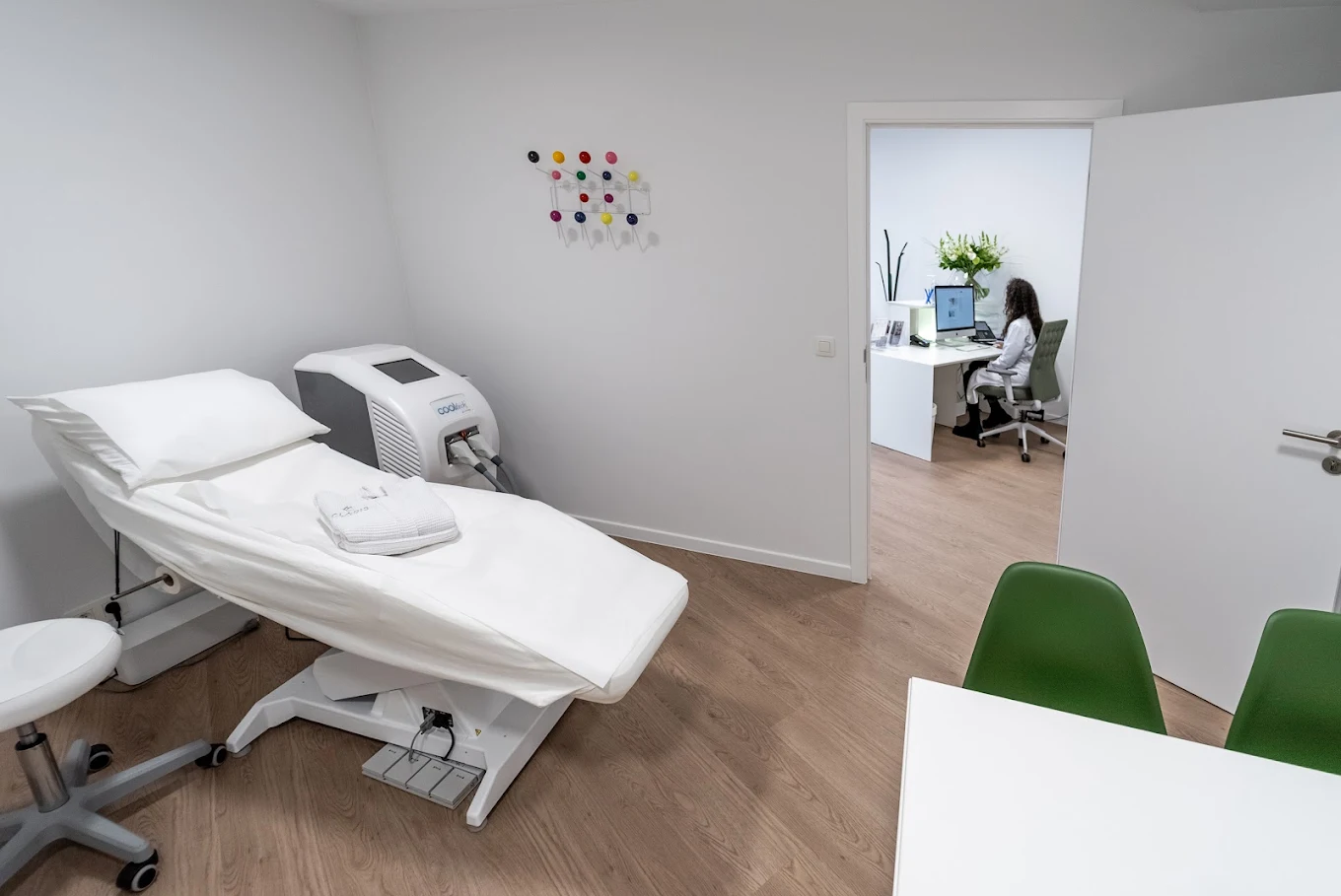 Une salle d'examen médical propre et moderne avec un lit réglable blanc, une serviette pliée, du matériel médical, des chaises vertes et une porte ouverte montrant une personne travaillant sur un ordinateur dans un bureau adjacent.