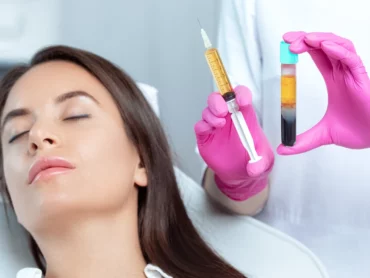 A woman with closed eyes is lying down while a person wearing pink gloves holds a syringe and a vial containing separated blood components, preparing for a medical or cosmetic procedure.