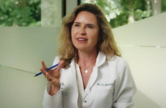 Une femme en blouse blanche tenant un stylo bleu prend la parole. Elle a de longs cheveux bruns ondulés et porte un collier en or. Le nom Dr. C. DeWULF est brodé sur sa blouse. Un fond vert flou est visible.