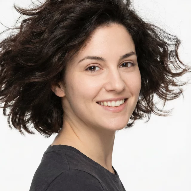 Une jeune femme aux cheveux bruns ondulés et à la peau claire sourit à l'appareil photo. Elle porte un t-shirt gris foncé et pose sur un fond blanc uni.