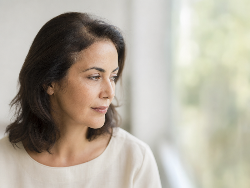 Une femme aux cheveux foncés, vêtue d'un haut beige clair, regarde vers sa gauche, l'air pensif. Elle se trouve à l'intérieur, avec en arrière-plan une fenêtre à travers laquelle on aperçoit un décor flou baigné de soleil.