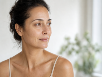 Une femme aux cheveux foncés attachés en arrière regarde sur le côté, le visage impassible. Elle porte un débardeur beige et se trouve à l'intérieur, baignée d'une douce lumière naturelle, avec une plante verte floue en arrière-plan.