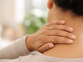 Une personne se tient la nuque d'une main, ce qui laisse supposer qu'elle ressent une gêne ou une douleur. On voit cette personne de dos, et l'arrière-plan est légèrement flou.