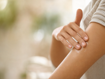 Une personne applique de la lotion sur son avant-bras avec les doigts. L'arrière-plan est légèrement flou, et la mise au point est faite sur la main et le bras. La personne porte une chemise à manches longues de couleur claire.