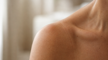 Gros plan sur l'épaule et la clavicule nues d'une personne, avec une marque de bronzage visible. Le teint est clair à moyen et l'arrière-plan est légèrement flou, dans des tons neutres.