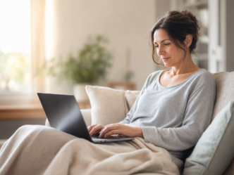 Une femme est assise sur un canapé, une couverture légère posée sur les jambes, et utilise un ordinateur portable. Elle porte un t-shirt gris à manches longues et semble concentrée. La scène se déroule dans un salon lumineux et chaleureux, avec des plantes en arrière-plan.