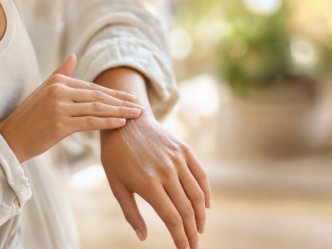 Une personne s'applique de la lotion sur le dos de la main. L'arrière-plan est légèrement flou, et la personne porte une chemise à manches longues de couleur claire.