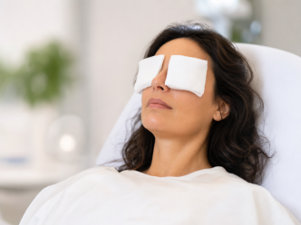 Une femme est allongée sur une table de soins, les yeux recouverts de deux compresses en tissu blanc. Elle est recouverte d'une couverture blanche et semble se trouver dans un spa ou un établissement médical. L'arrière-plan est flou.