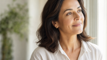 Une femme aux cheveux foncés mi-longs, vêtue d'un chemisier blanc, regarde par la fenêtre avec un léger sourire. Une douce lumière naturelle illumine son visage. On aperçoit, en arrière-plan, une plante verte floue.