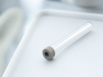Un tube à essai en plastique transparent ou en verre, muni d'un bouchon en caoutchouc gris, repose sur un plateau blanc dans un environnement très éclairé.