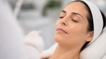 Une femme aux cheveux foncés, portant un bandeau blanc, est allongée sur une table de soins, les yeux fermés, vêtue d'un peignoir blanc, dans ce qui semble être un spa ou un centre de soins de la peau.