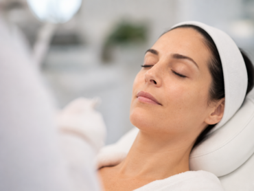 Une femme aux cheveux foncés, portant un bandeau blanc, est allongée sur une table de soins, les yeux fermés, vêtue d'un peignoir blanc, dans ce qui semble être un spa ou un centre de soins de la peau.