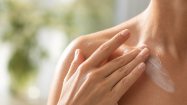 Une personne applique du bout des doigts une couche de lotion ou de crème blanche sur son épaule nue et le haut de sa poitrine. L'arrière-plan est légèrement flou et on aperçoit de la verdure.
