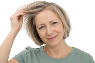 Une femme d'âge mûr, aux cheveux blonds courts, vêtue d'un t-shirt vert, se touche les cheveux et esquisse un léger sourire. Elle regarde directement l'objectif, devant un fond uni et clair.